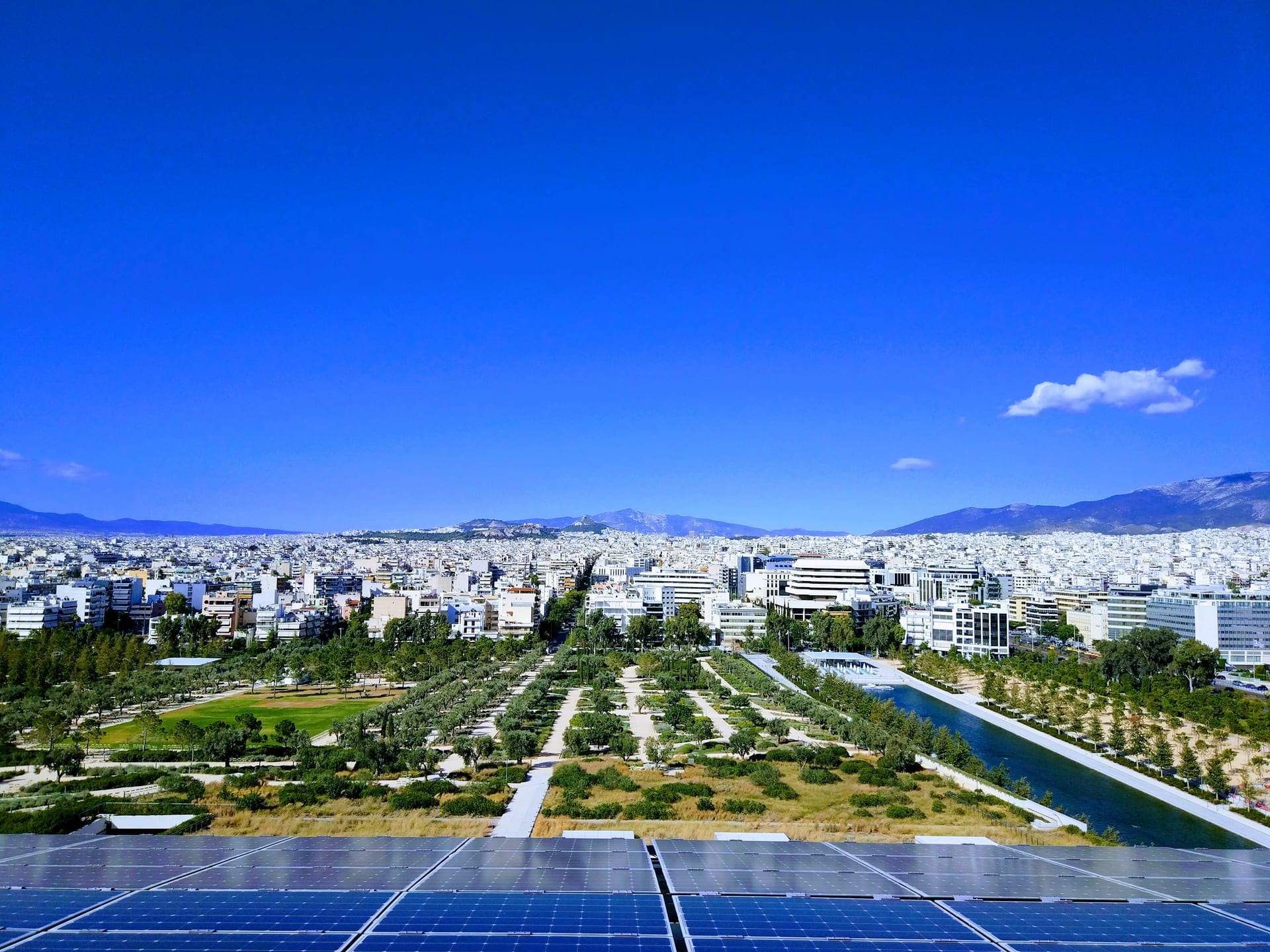View over Stavros Niarchos Park and its long canal from the solar-panel canopy of the Stavros Niarchos Foundation Cultural Center, with central Athens and the Acropolis in the distance.